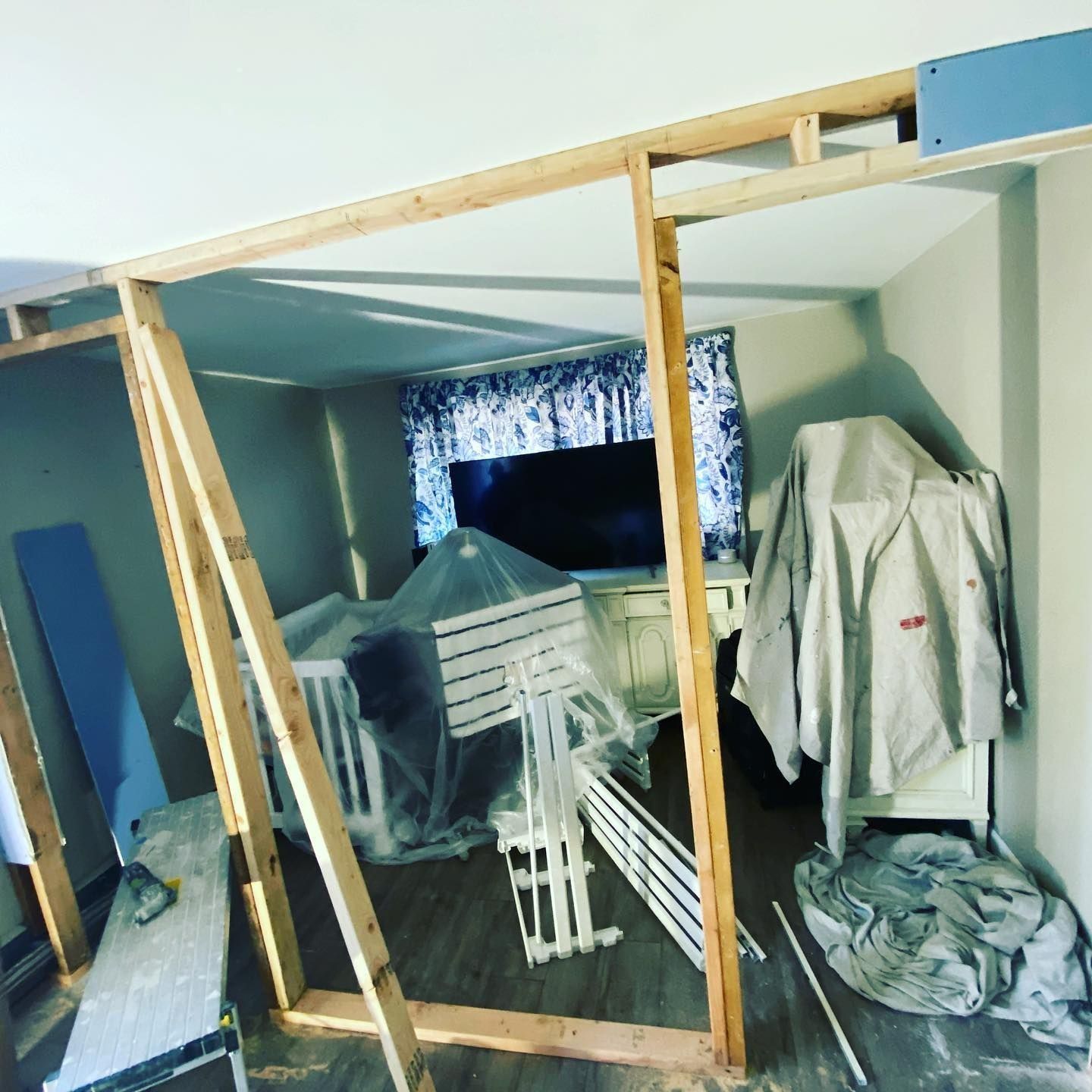 Room under construction with wooden frame dividing space; cribs covered in plastic, curtains visible in the background.