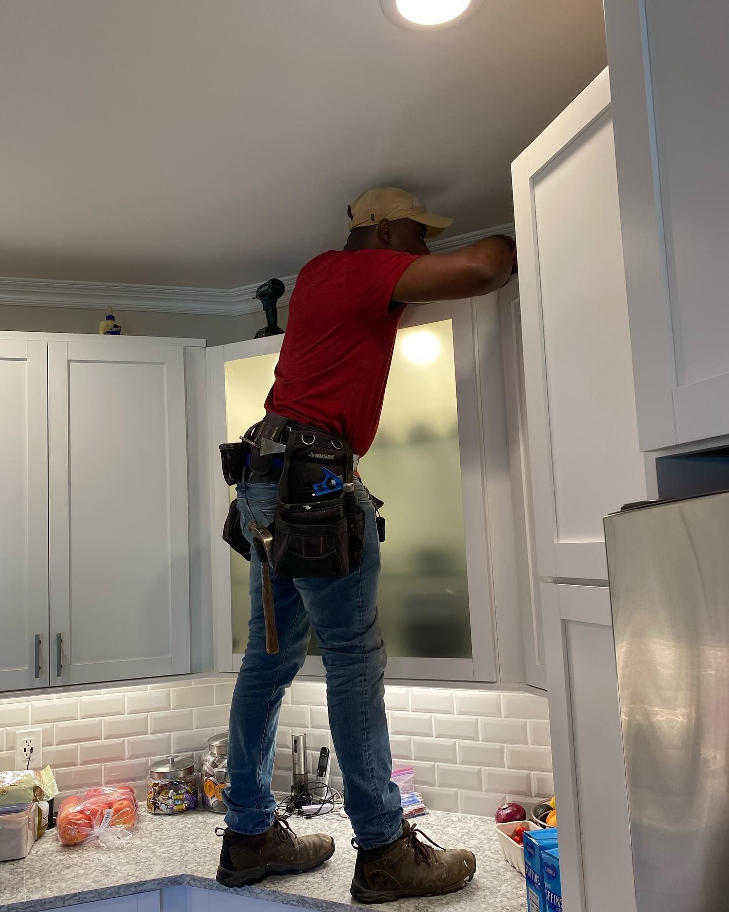 Person in red shirt and jeans installing a white cabinet above a countertop in a kitchen.