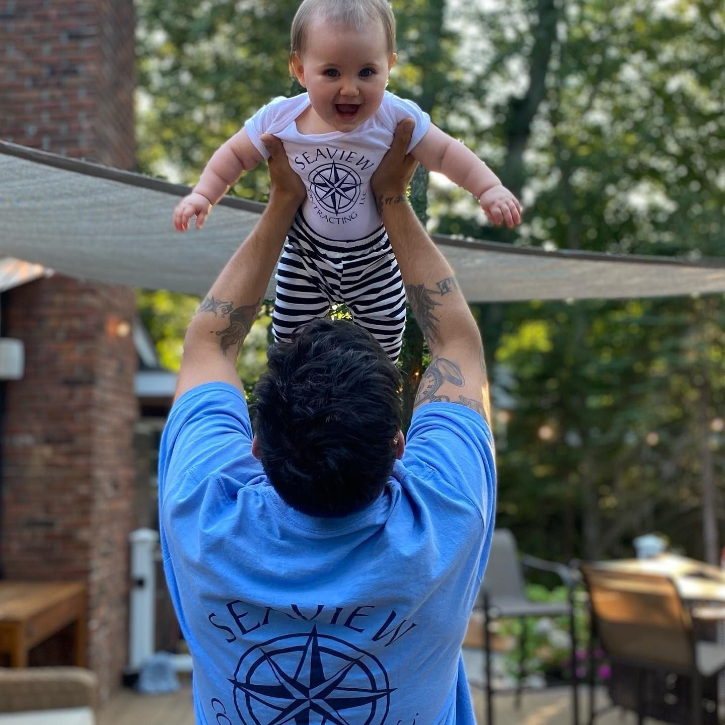 Man holding up baby; outdoors. The baby smiles, wearing a white onesie and striped pants. The man wears a blue shirt.