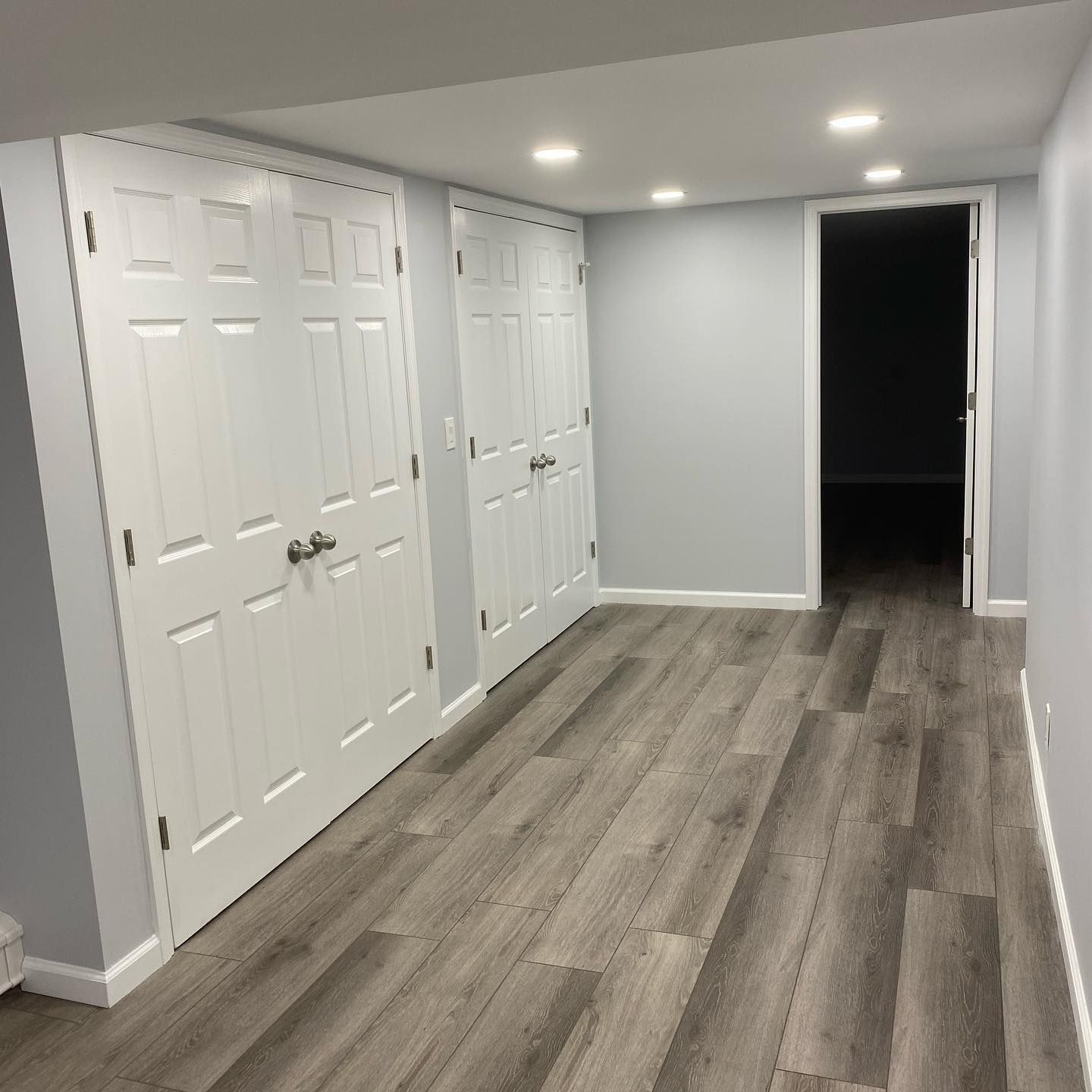 A narrow hallway with gray wood-look flooring, white doors, and a doorway. The walls are light gray.