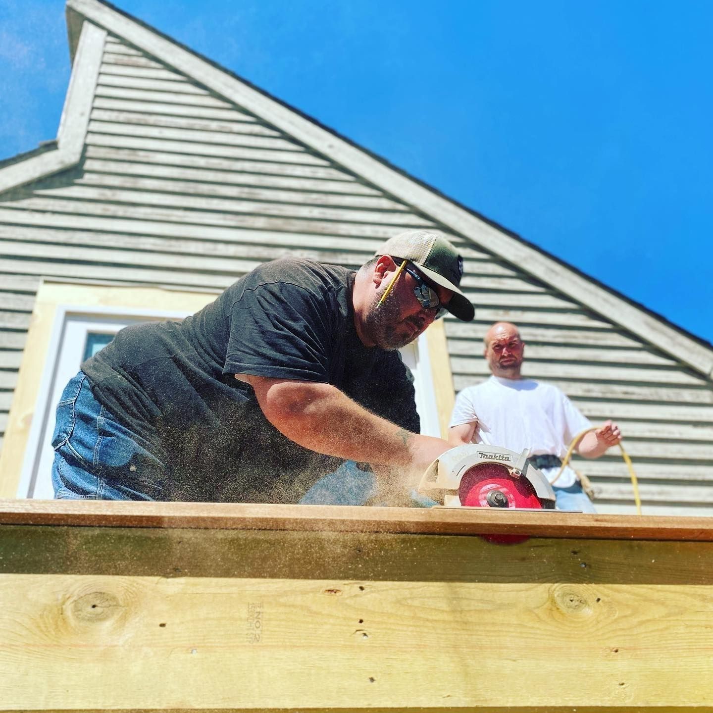 Man sawing wood with a circular saw on a deck. Another man watches in background. Blue sky overhead.