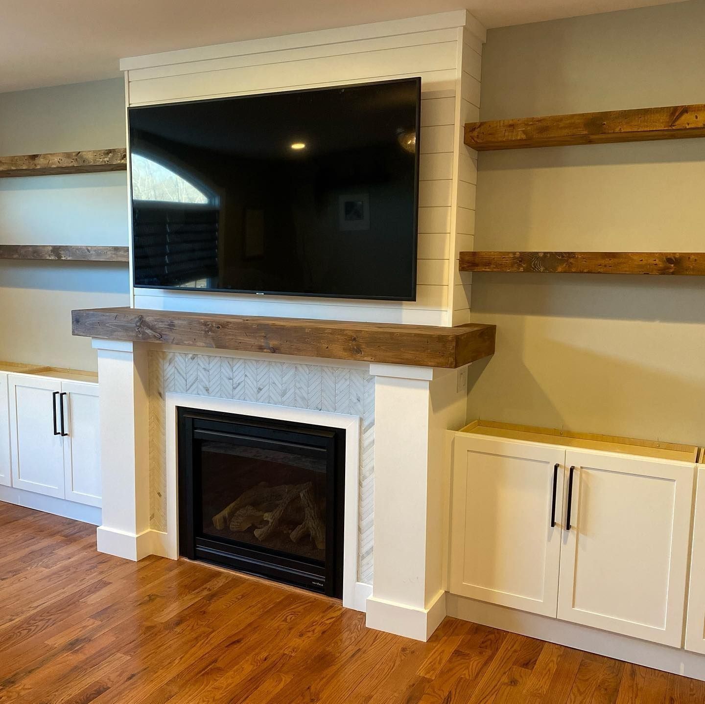 Fireplace with large TV, wooden shelves, and white cabinets. Brown wooden mantel and trim.