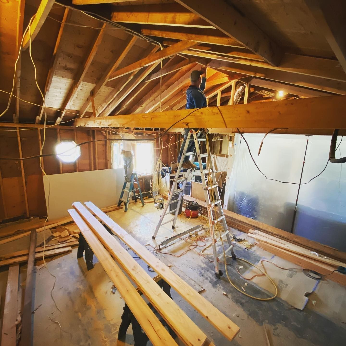 Construction crew working on an attic; exposed beams, ladders, drywall, and wooden planks.