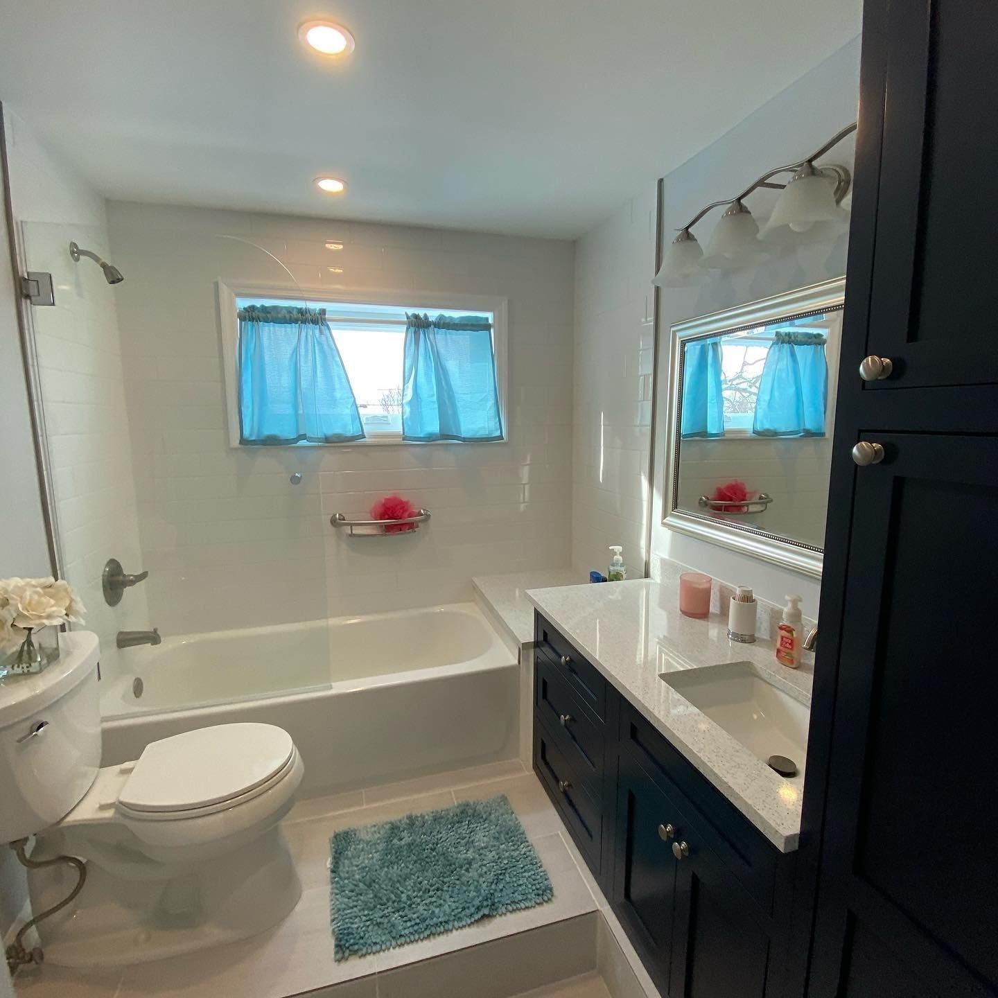 Bathroom with blue vanity, tub, and curtains; white walls, and a teal rug.