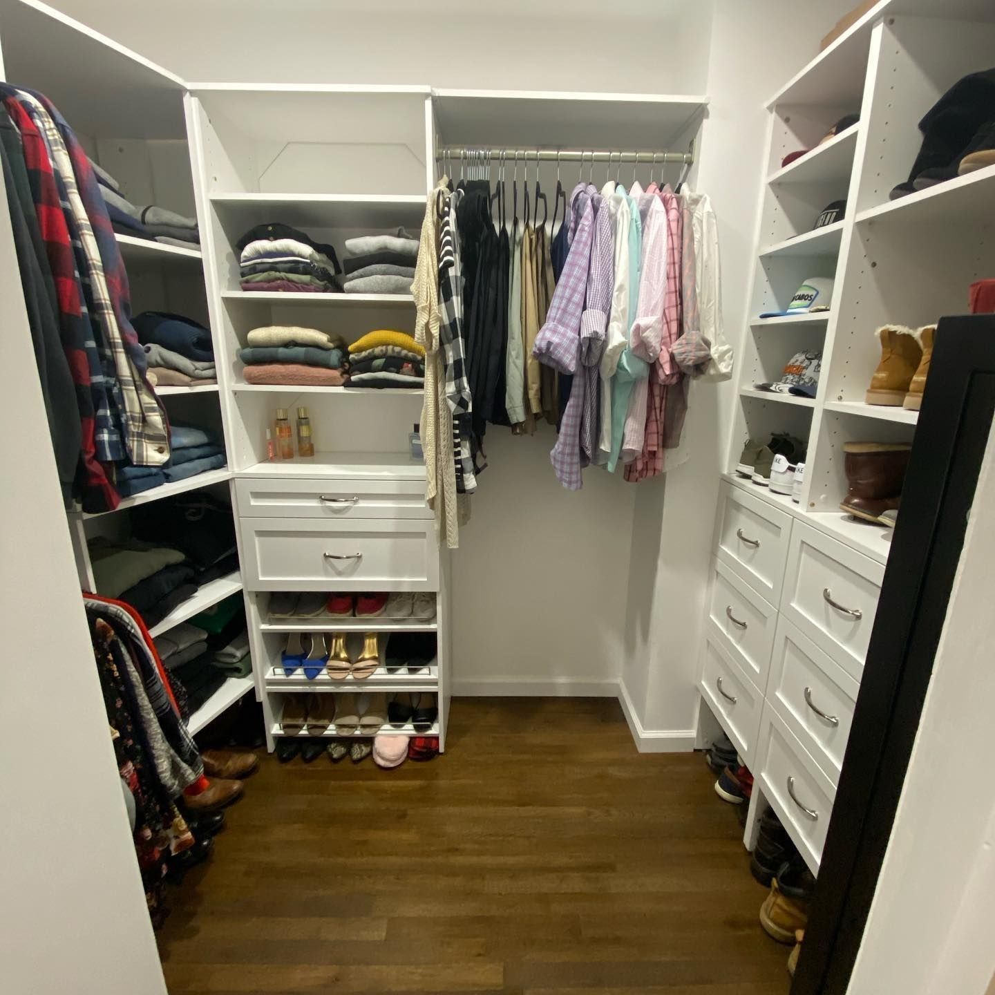 White walk-in closet with shelves and hanging rods; clothes and shoes are organized on brown wood floor.