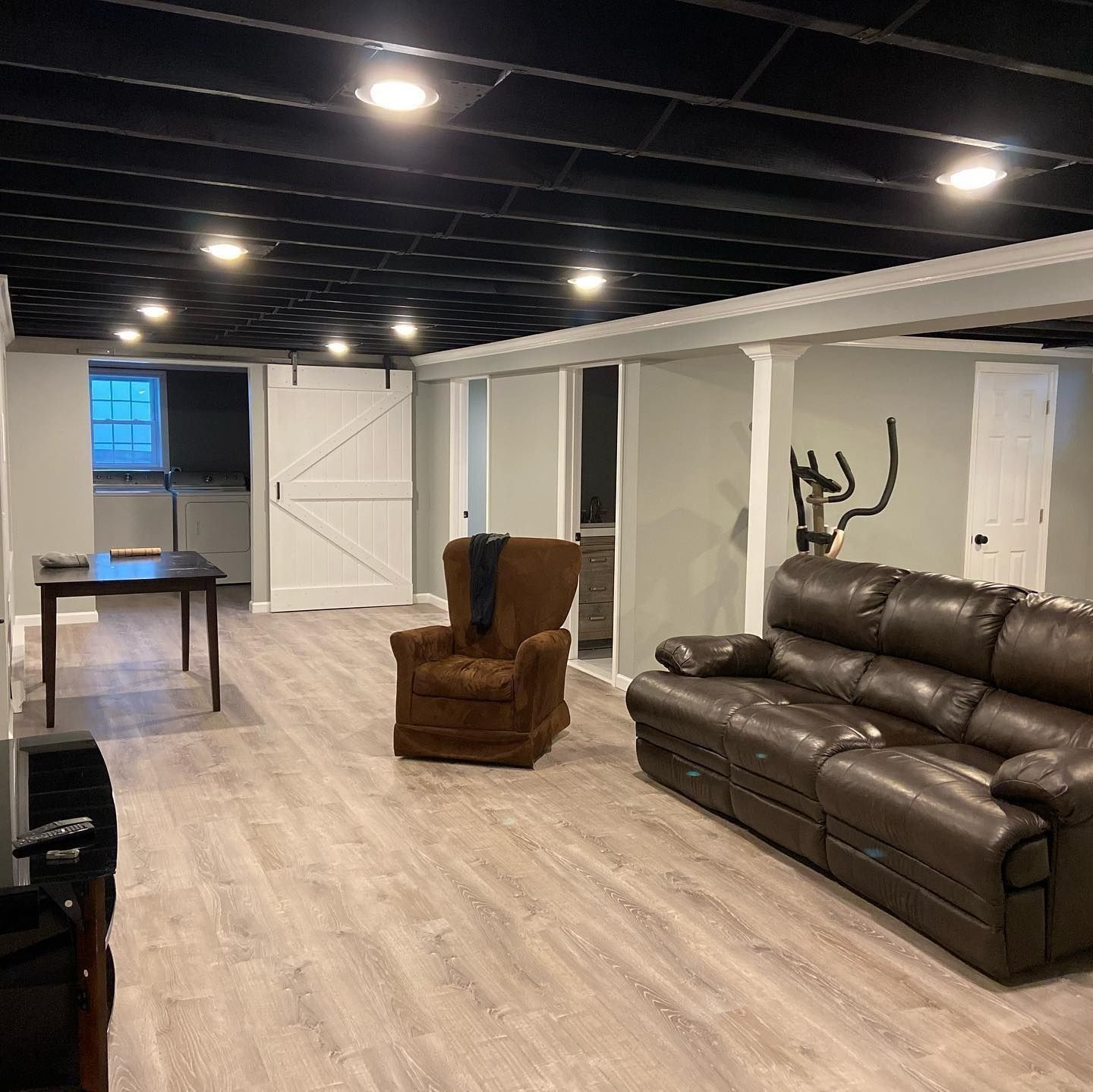 Finished basement with brown leather sofa, chair, and black ceiling. Light gray walls, light flooring.