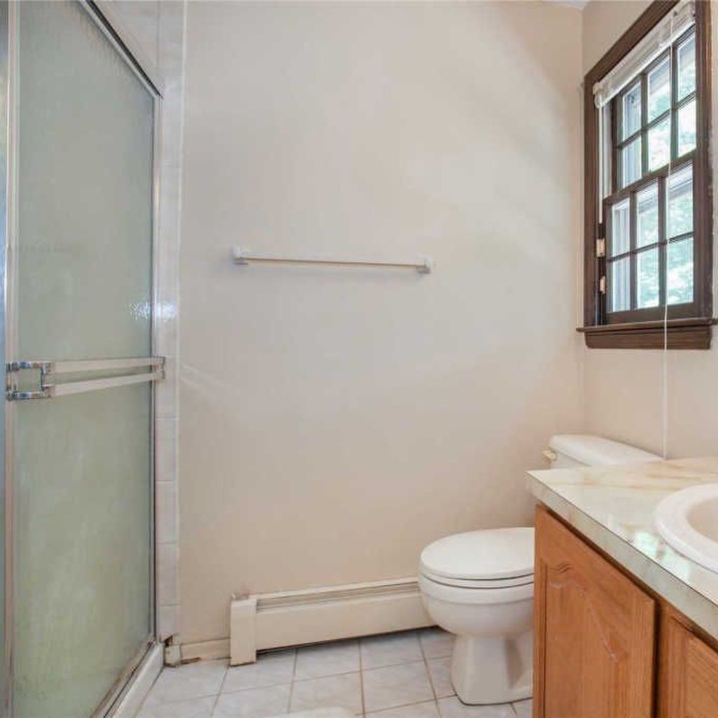 Bathroom with shower, toilet, window. Beige walls, wood cabinet, white fixtures, and light blue tile flooring.