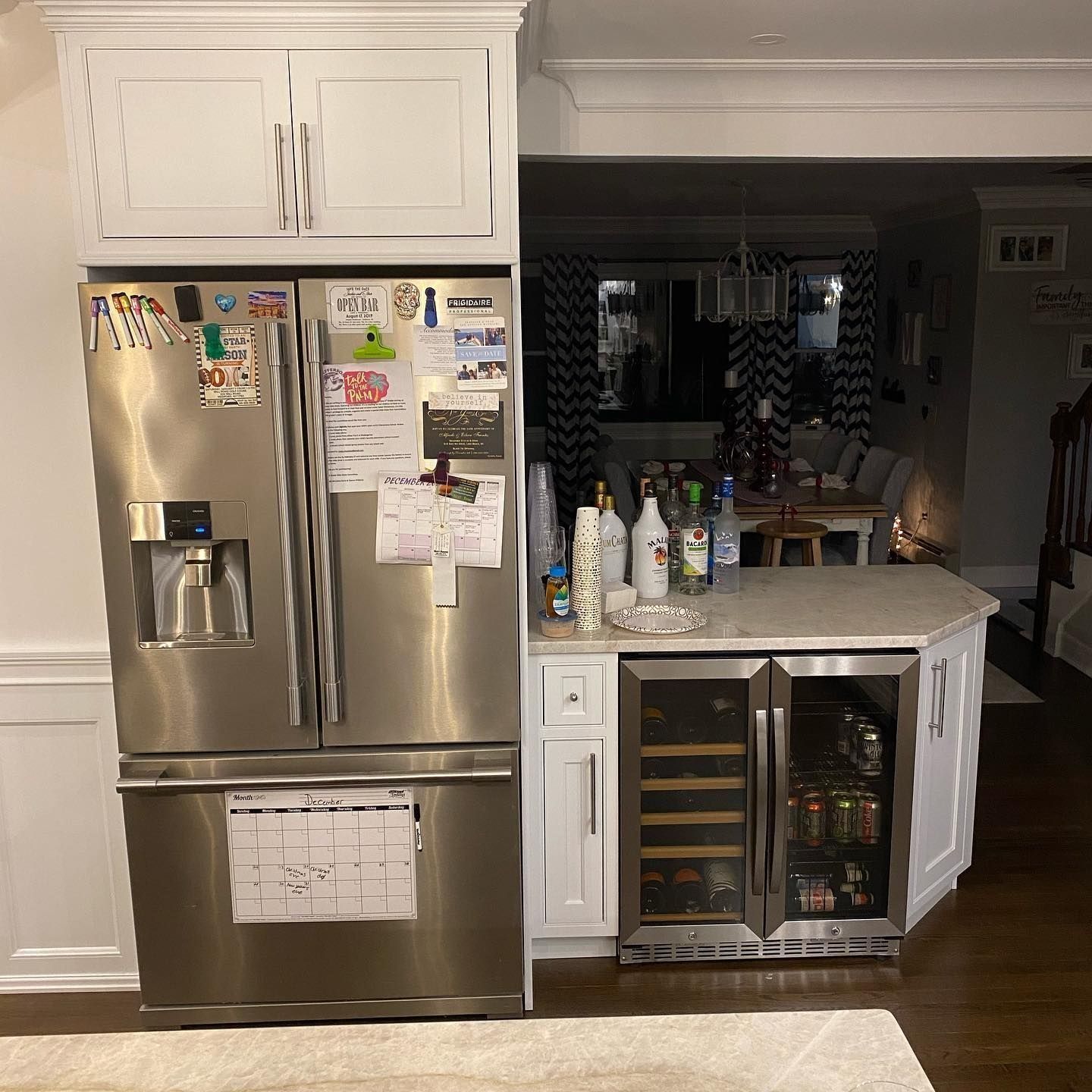 Refrigerator next to a built-in bar area with a wine cooler. White cabinets and a dark dining room visible.