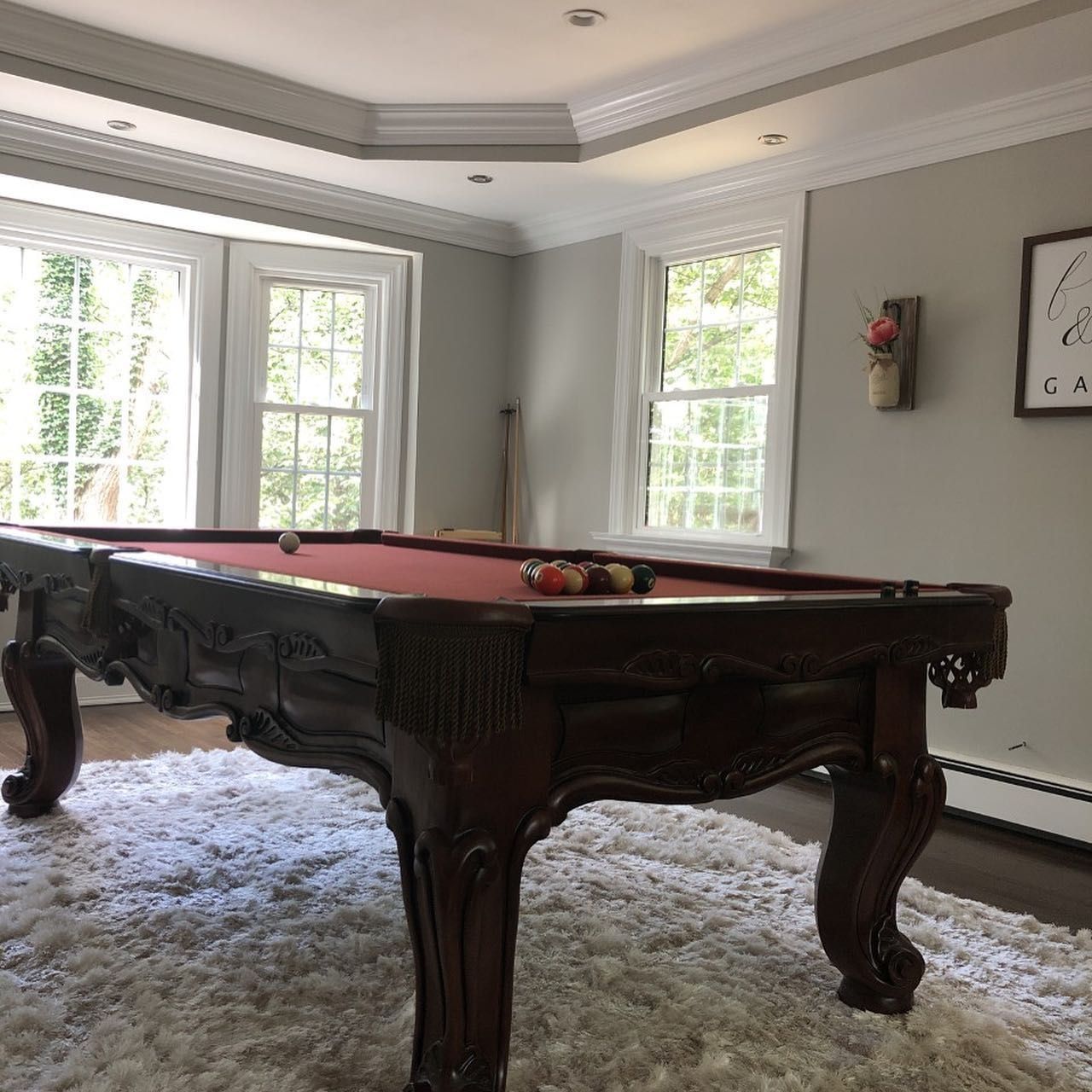 A pool table with red felt sits on a fluffy white rug in a well-lit room with windows.