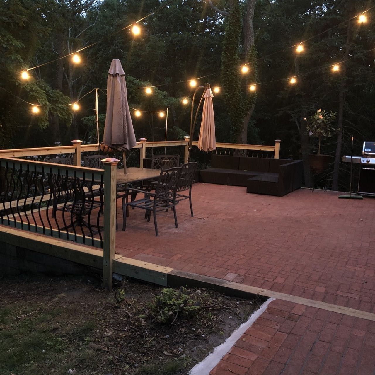 Patio with string lights, brick floor, tables, chairs, and a dark wicker sofa under open umbrellas at dusk.