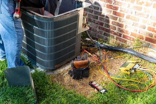 Technician working on outdoor HVAC unit beside brick wall, with tools and coiled wires on the grass.