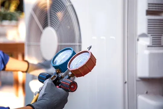 Gloved hand holding HVAC gauges beside an outdoor AC unit and fan