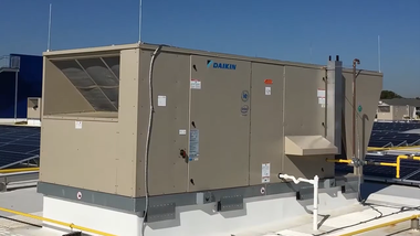A tan commercial HVAC rooftop unit sits on a white curb against a clear blue sky on a flat building roof.