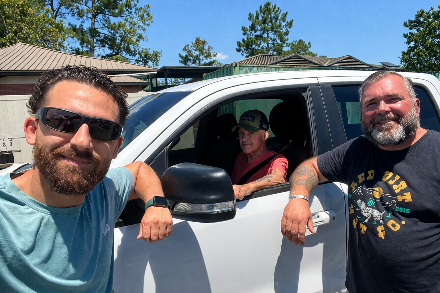 Two men stand smiling outside a white pickup truck, while a third person sits in the driver’s seat on a sunny day.