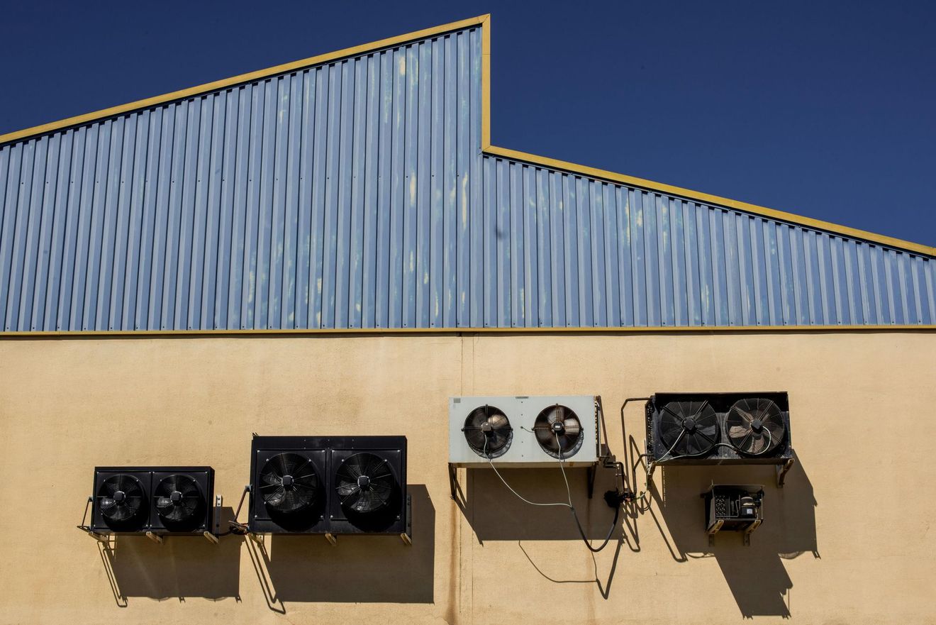 Four outdoor air conditioning units mounted on a beige wall beneath a blue corrugated metal roof.