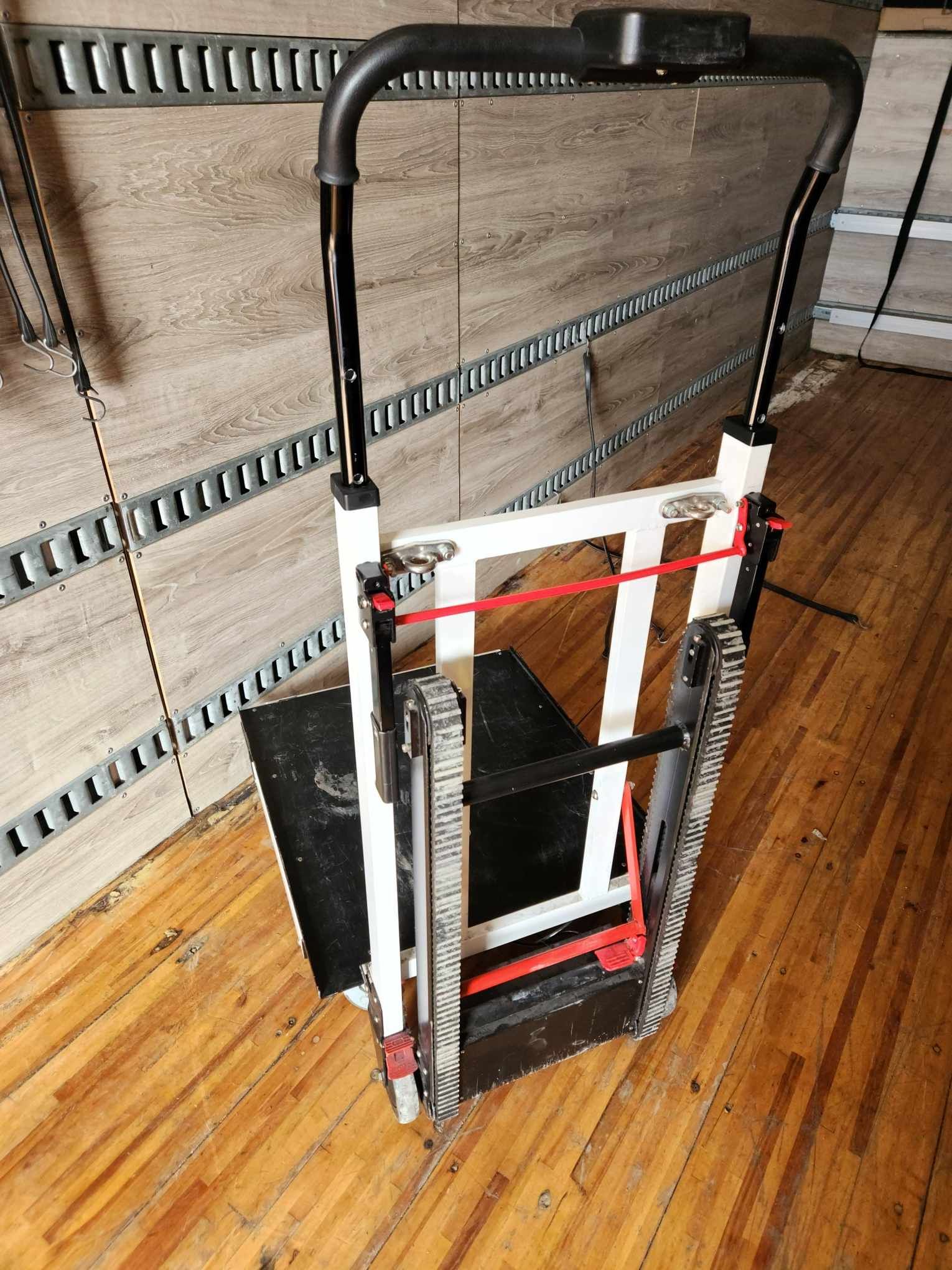 Un diable pour escaliers, cadre blanc, plateforme noire, élastique rouge. Appuyé contre un mur dans une pièce.