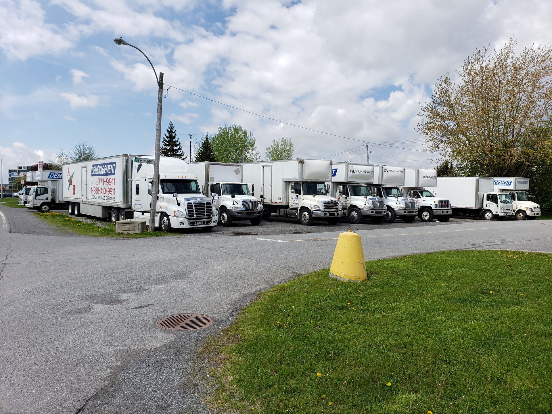 Des camions stationnés sur un terrain gravillonné avec une zone herbeuse et un lampadaire sous un ciel nuageux.
