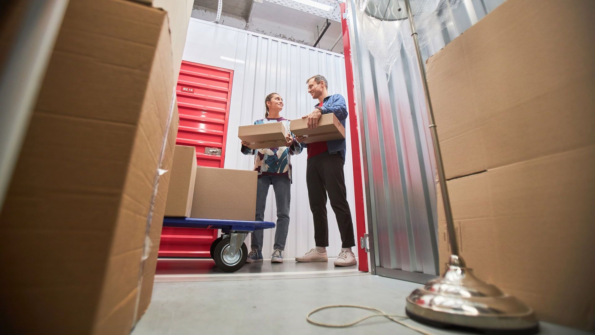 Deux personnes dans un garde-meuble transportant des cartons. Chariot et lampe visibles.