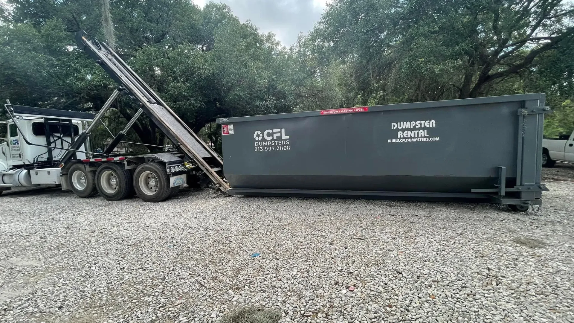 A dumpster is being loaded onto a truck in a gravel lot.