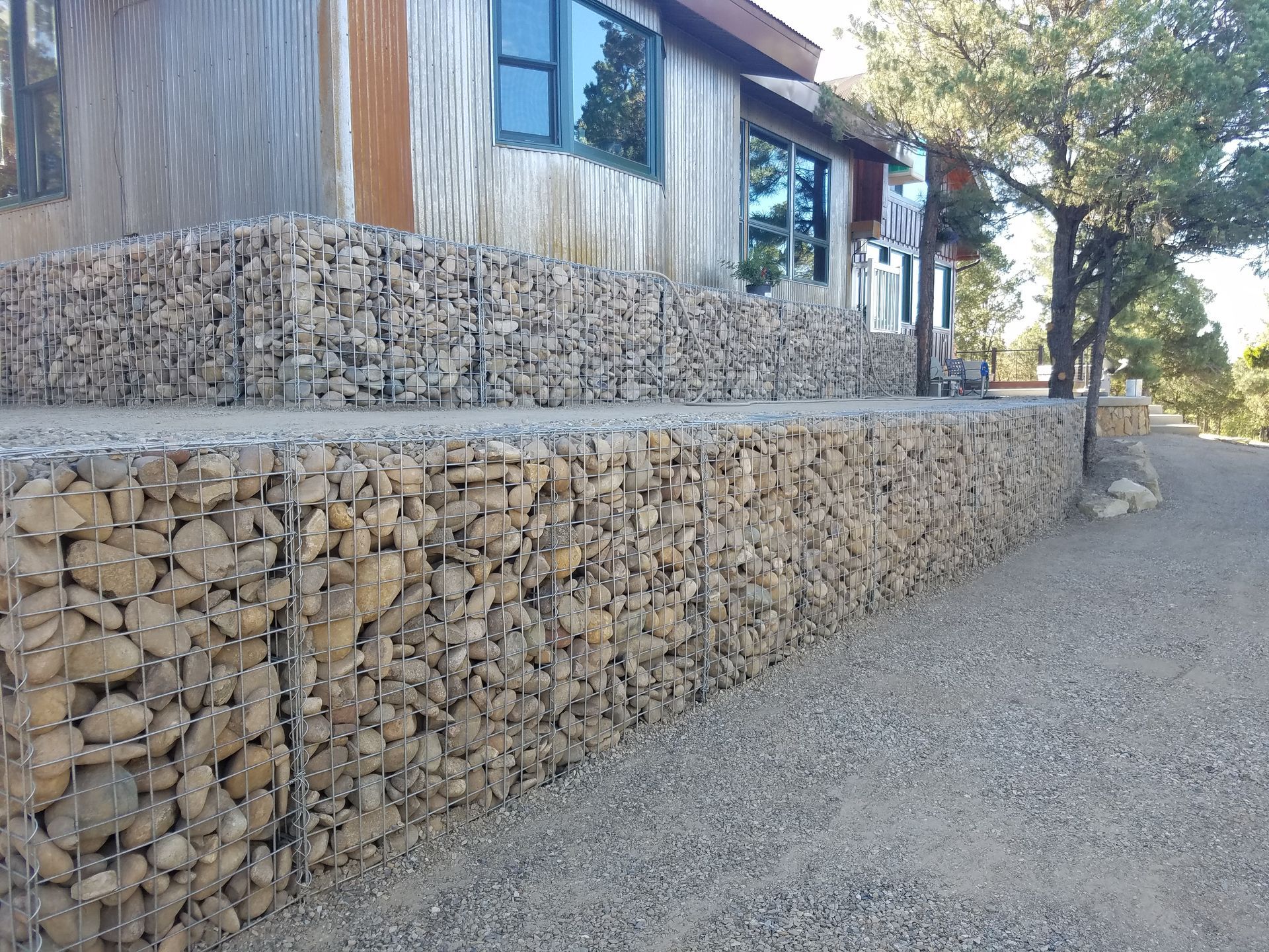 A house with exterior walls and a raised patio supported by layered, wire-mesh gabion retaining walls filled with rocks.