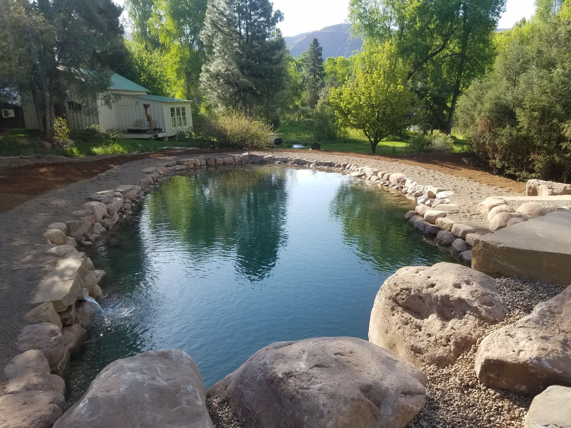 A serene pond with a stone border, a small waterfall, and a house in the background, set amidst lush green trees.