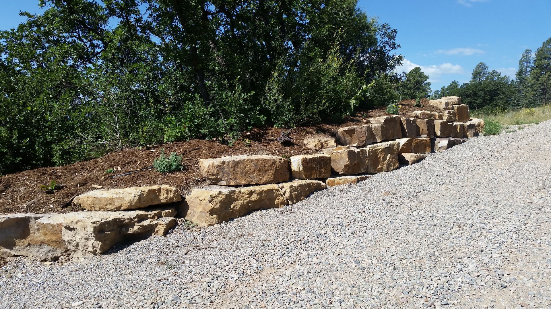 A tiered retaining wall made of large, tan, rectangular stones next to a gravel driveway and a mulch-covered slope.