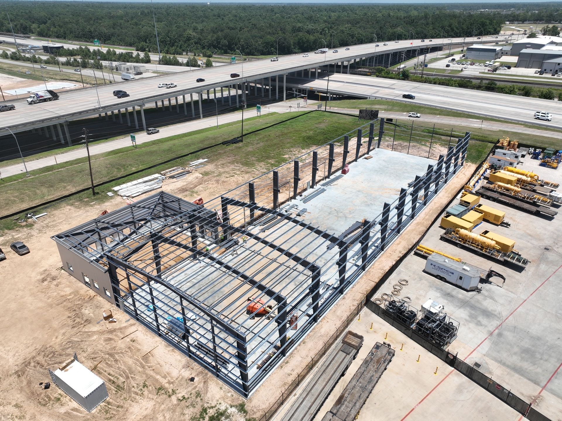 Construction site with steel frame building under construction, heavy equipment, and workers. Brown earth, blue sky.
