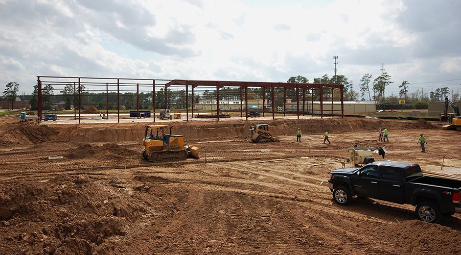 Construction site with steel frame building under construction, heavy equipment, and workers. Brown earth, blue sky.