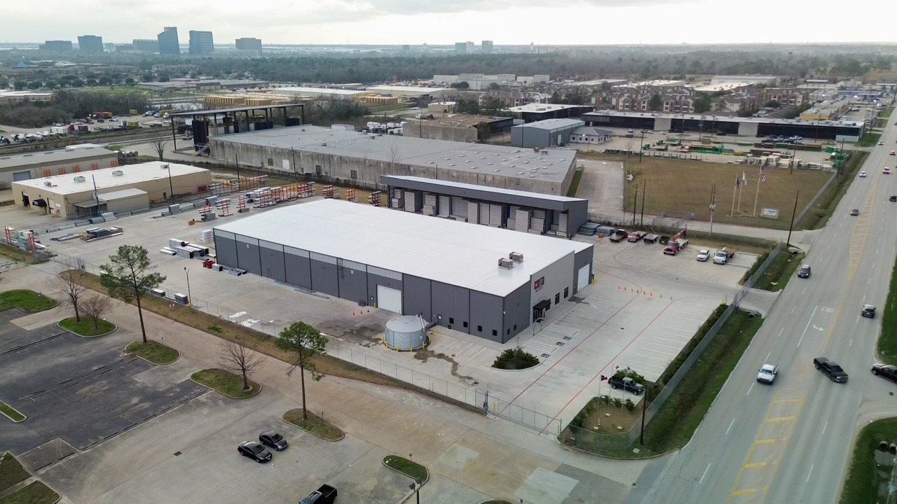 Aerial view of industrial buildings with a gray facade, adjacent to a road with traffic, and a city skyline in the distance.