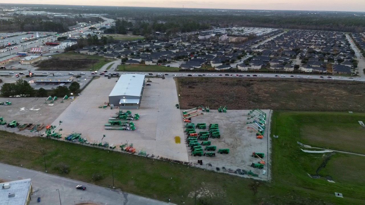 Aerial view of a commercial lot with a large building, various green equipment, and residential housing in the background.