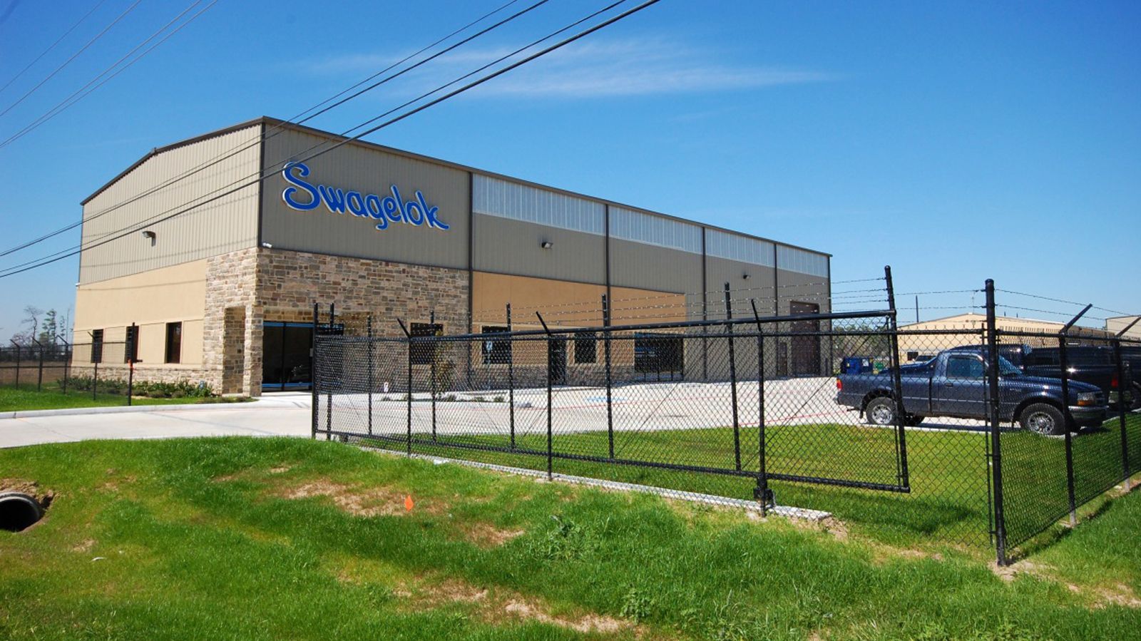 Swagelok building exterior, light brown and gray with black fence, blue sky, truck parked outside.