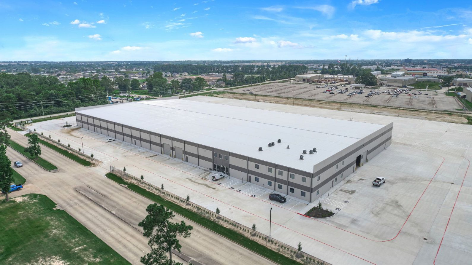 Aerial view of a large, light-colored industrial warehouse with a flat roof; surrounded by a concrete lot.