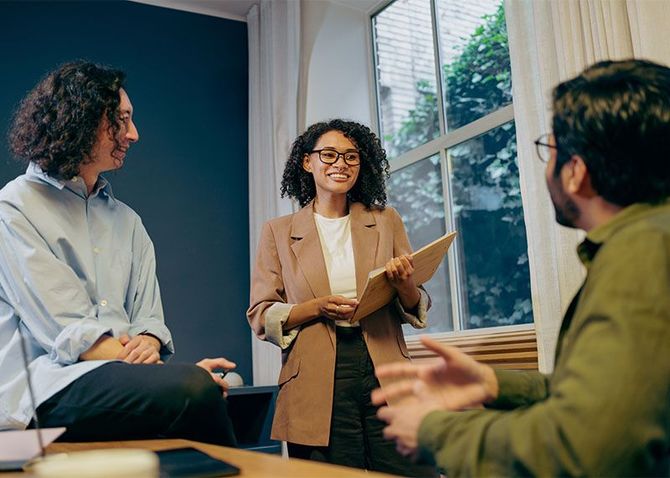 Three people in a meeting, one speaking and holding a notepad by a window in a bright office