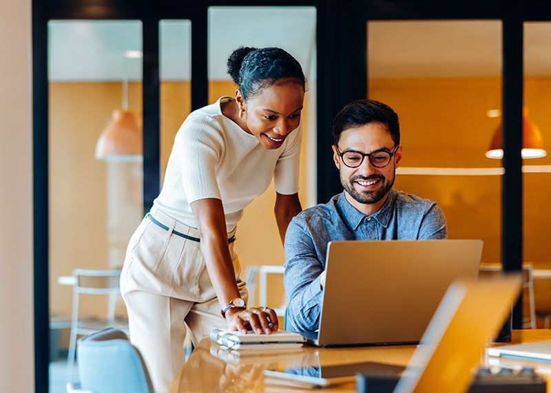 Two coworkers smiling at a laptop in a modern office, with one leaning over to point at the screen.