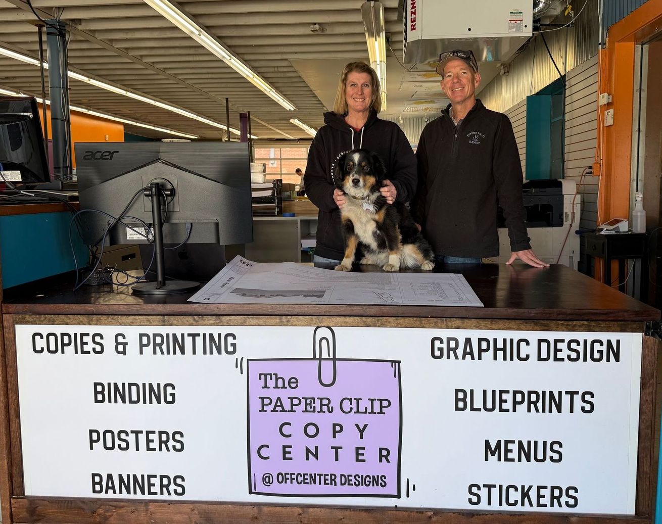 A man and woman are standing in front of a counter at the paper clip copy center holding a dog.