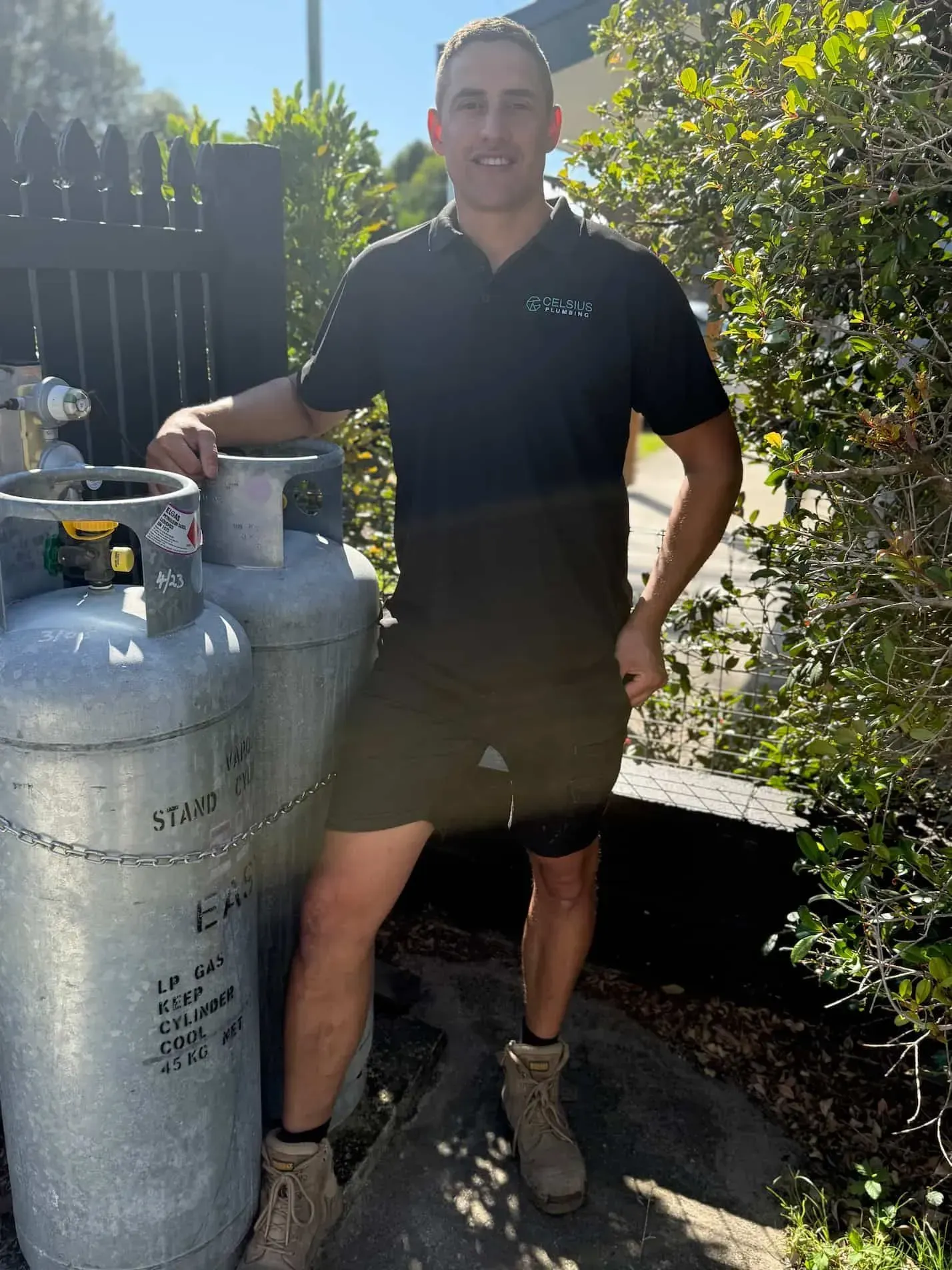 Man in black polo shirt and shorts stands next to two propane tanks outdoors.