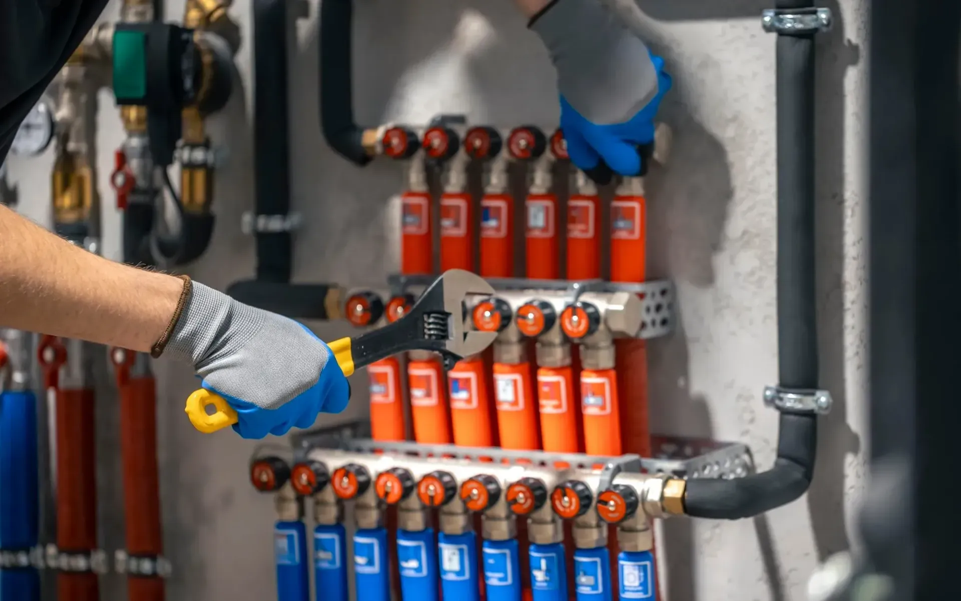 Plumber in gloves using a wrench on a manifold system with orange and blue pipes.