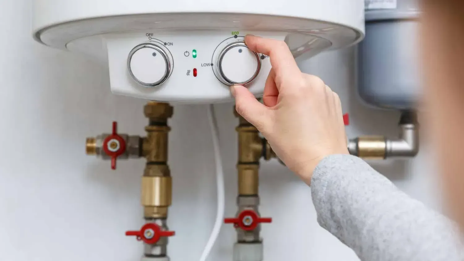 A person's hand turning a knob on a white water heater, next to copper pipes and red valves.