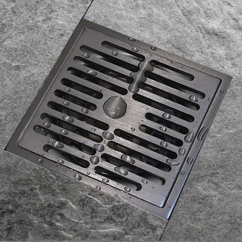 Square metal drain cover with water droplets on gray tiled floor.