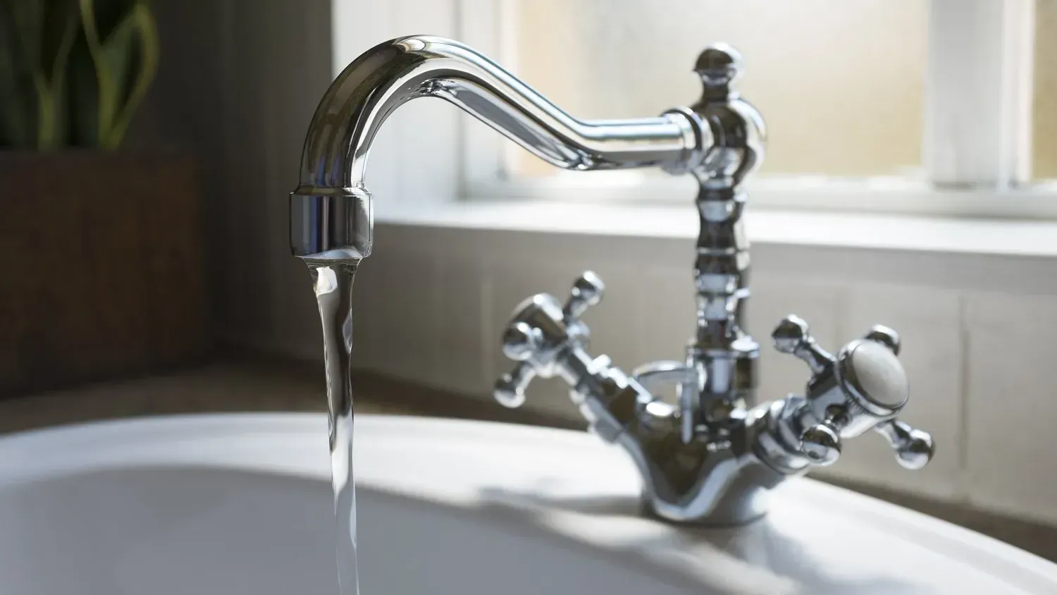 Chrome faucet with running water over a white sink, near a window.