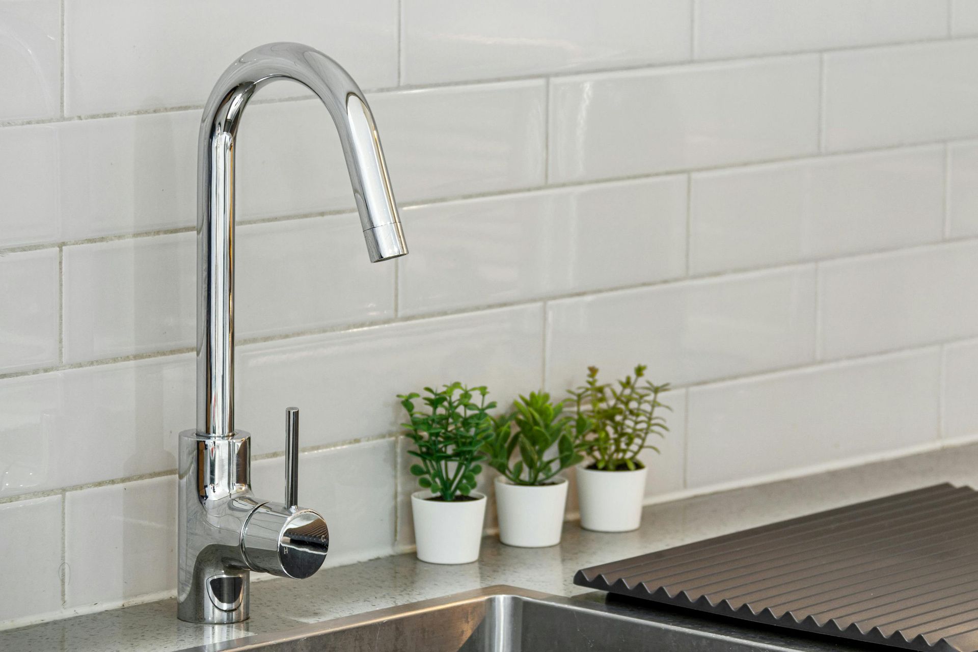 Chrome kitchen faucet beside small potted plants on a counter against white tile backsplash