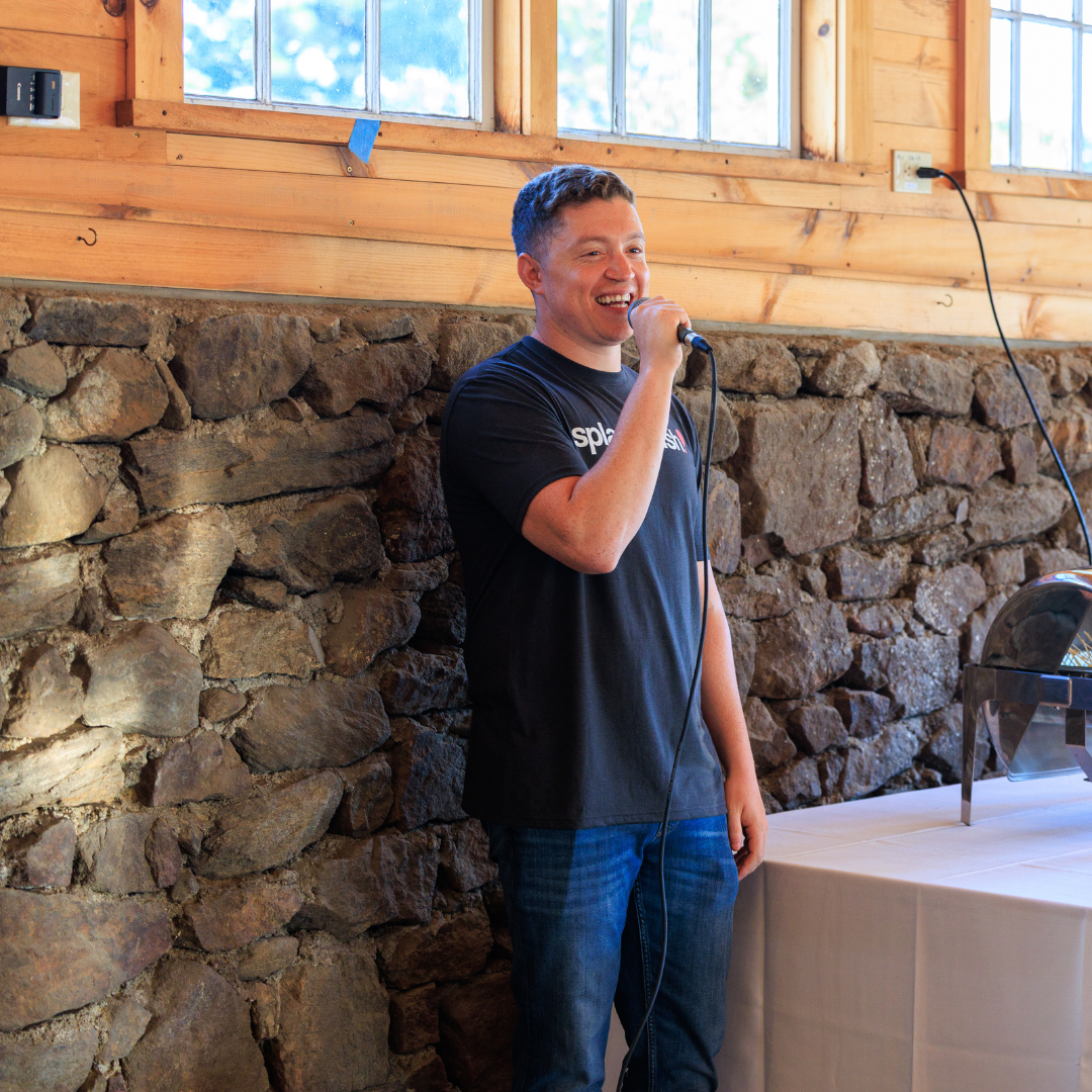 Man speaking into a microphone near a stone wall, wearing a black t-shirt and jeans.