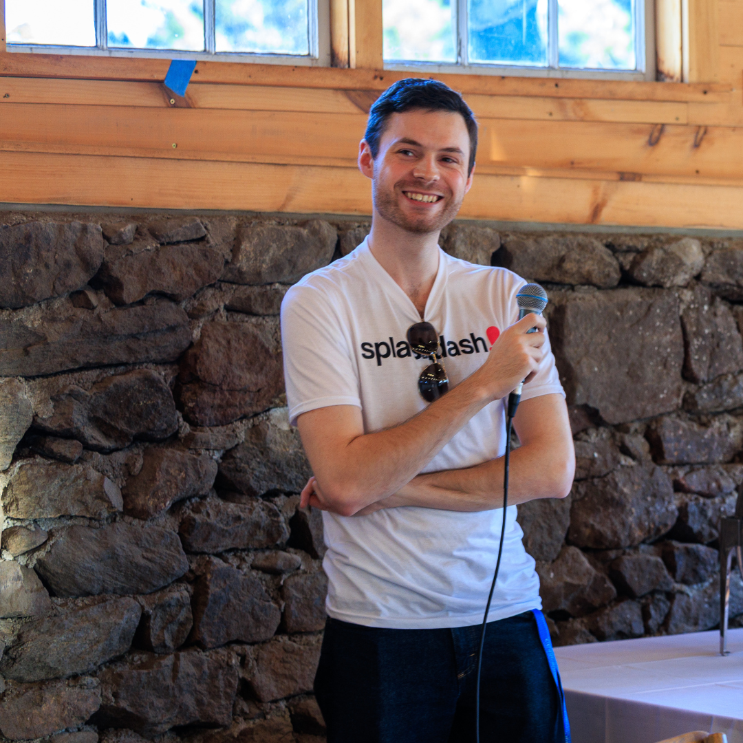 Man smiling, holding a microphone, standing in front of a stone wall and a window.