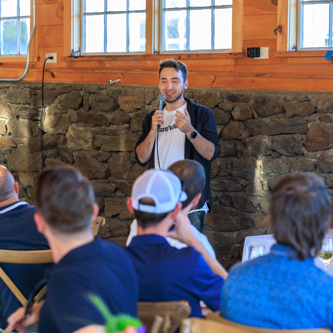 Man speaking into a microphone in front of a group, wood-paneled room with stone wall backdrop.