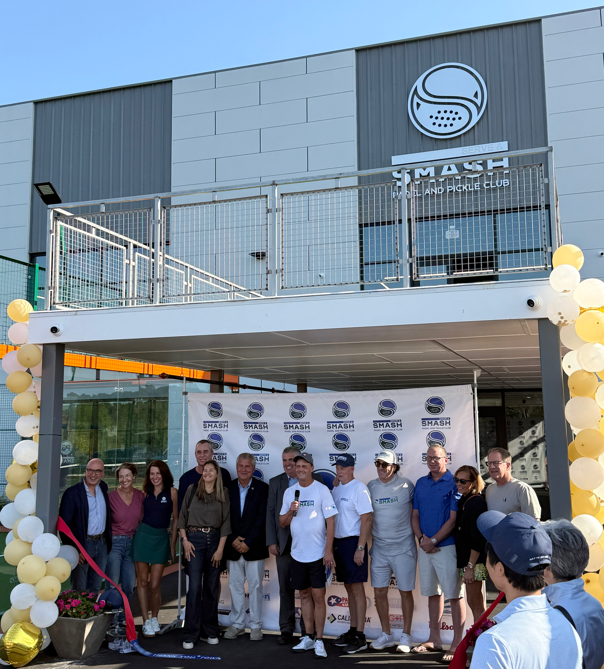 Ribbon-cutting ceremony at new sports facility. People stand in front of building with balloons. Sign above the entrance.