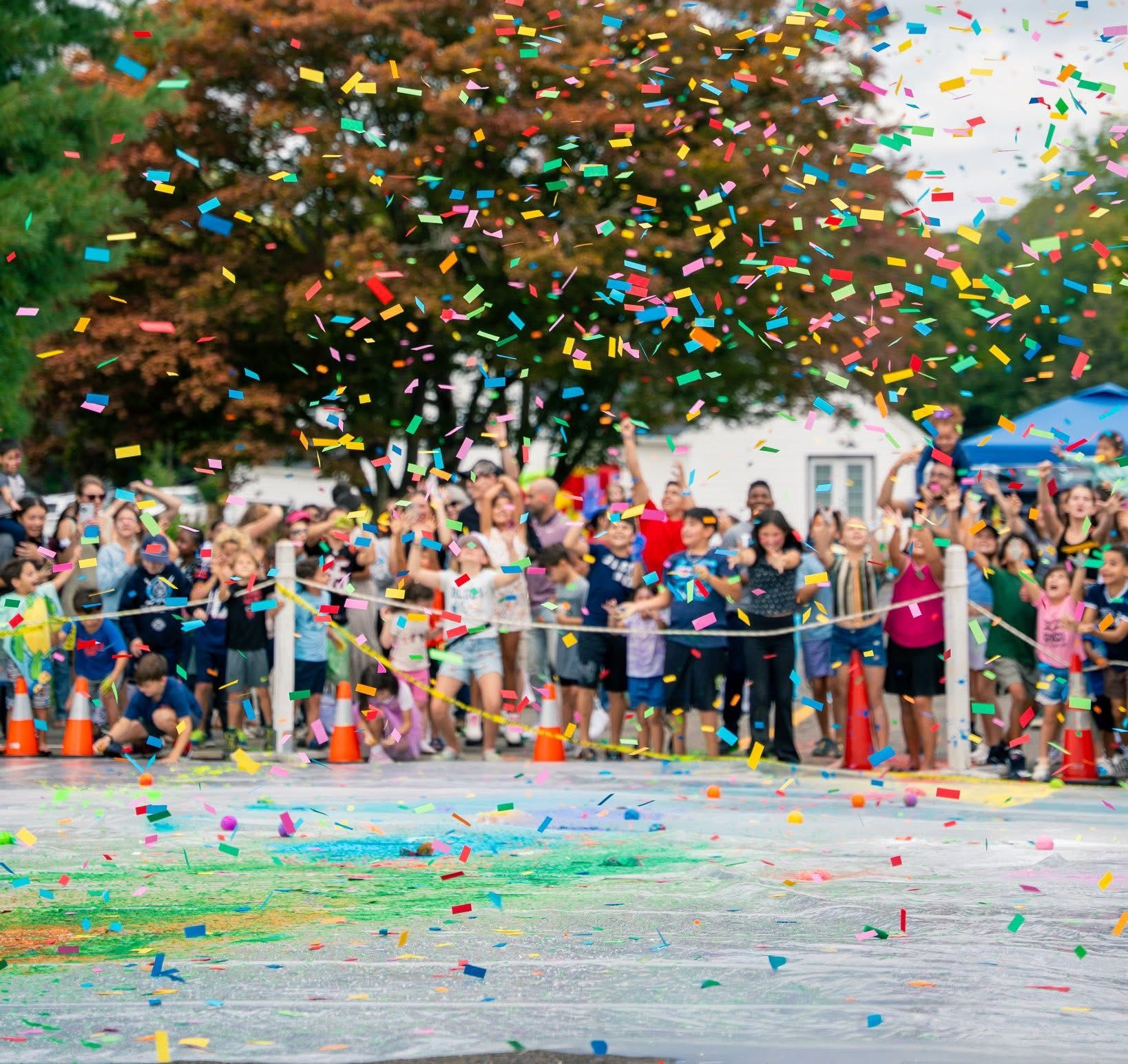 Crowd cheers as colorful confetti rains down during an outdoor event; people of all ages gathered.