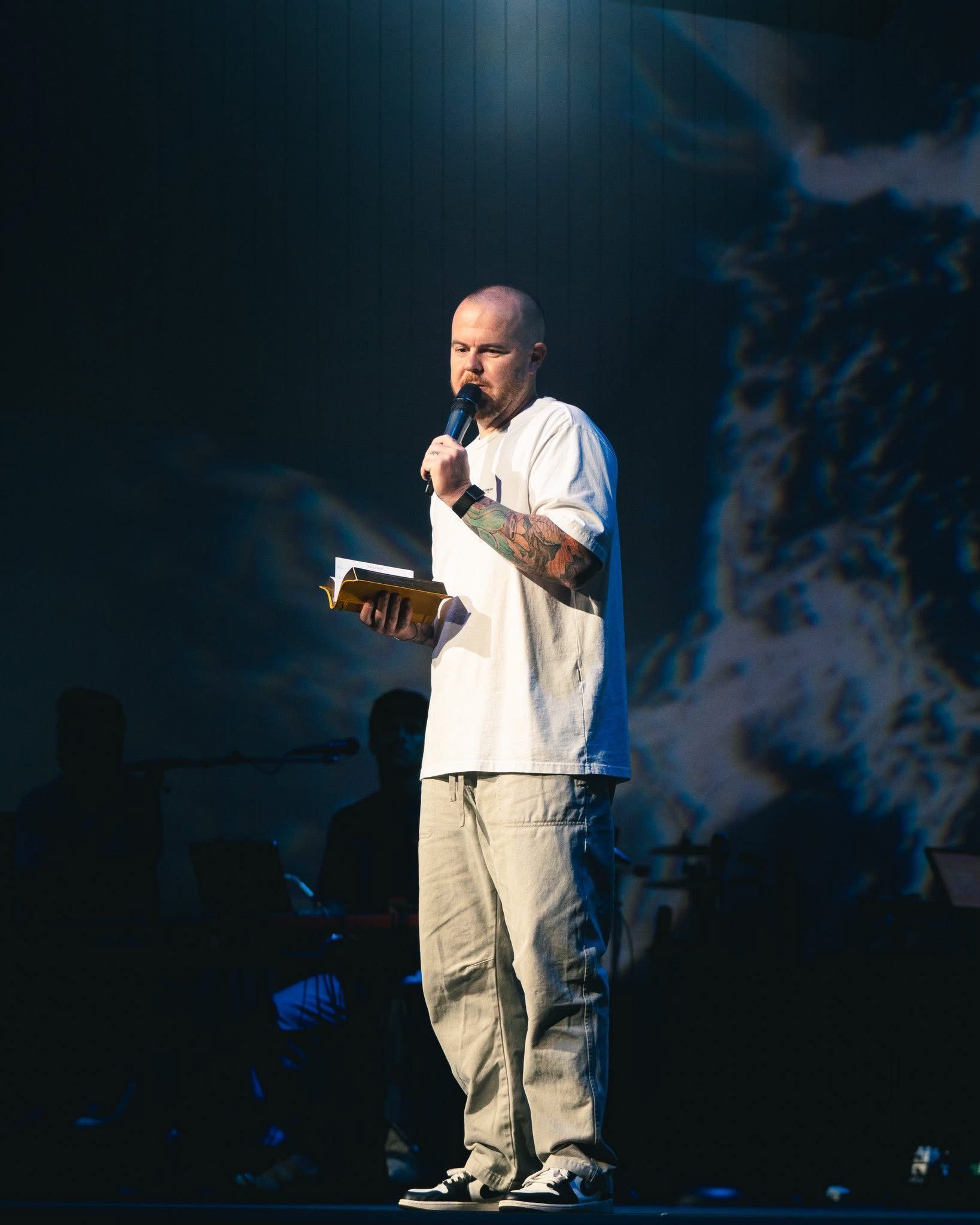 Man with tattoos holding a microphone and book, speaking on stage with a cloudy backdrop.