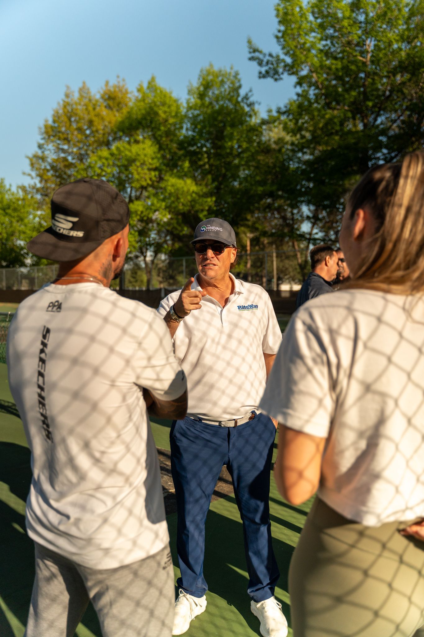 Group of people talking near a chain-link fence, outdoors in a park, backlit by sunlight.