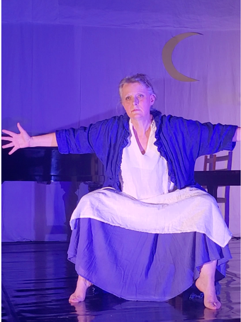 Femme en costume bleu et blanc, bras ouverts, accroupie sur scène sous un croissant de lune.