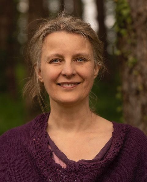 Femme souriante, vêtue d'un châle violet, se détachant sur un fond de verdure floue.